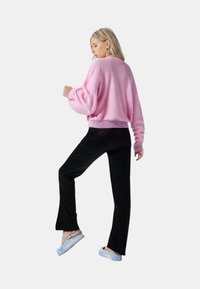 Maglione lavorato a maglia di un rosa chiaro con maniche lunghe e orlo a coste, abbinato a pantaloni neri. Modello che indossa scarpe slip-on blu con motivo.