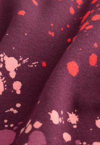 Violetter Stoff mit unregelmäßigen rosa und roten Farbspritzmustern, die Textur und Falten im Material zeigen.