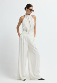 Witte haltertop en wijde broek outfit. Glad materiaal, geplooid ontwerp, gouden knoopdetails, gecombineerd met donkere zonnebril en puntige schoenen.