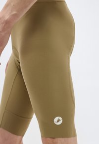 Közeli kép egy személyről, aki szoros olívazöld kerékpáros rövidnadrágot visel, amelyen a combon egy kis kör alakú skorpió logó látható.