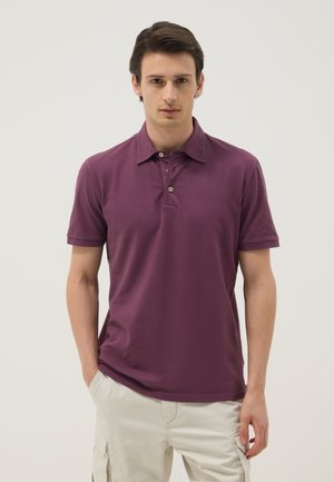 Junger Mann mit dunklen Haaren, der ein violettes Poloshirt und hellbeige Cargohosen trägt, steht vor einem schlichten hellen Hintergrund.