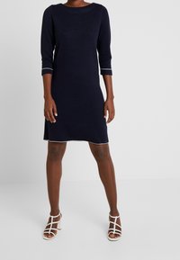 Robe en maille bleu marine avec manches trois-quarts et coutures contrastantes. Coupe droite avec un col arrondi. Associée à des sandales à talons blanches à brides.