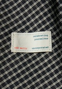 Tengerészkék és krémszínű kockás szövet háttér egy téglalap alakú fehér címkével. A szöveg így szól: „odd molly”, „önmagatok ünneplése”, „uncorporated”.