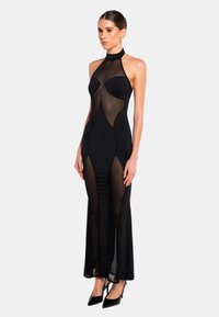 Svart halterneck-klänning med en figurnära bodice, genomskinliga paneler, geometriska utskärningar och en lång kjol med en sidoklyvning. Ackompanjeras av högklackade skor.