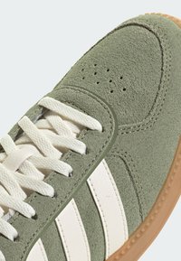 Zeleni antilop tenisica s bijelim kožnim prugama, teksturiranim gornjim dijelom, okruglim vrhom i ravnim bijelim vezicama. Gumeni potplat u boji gume.