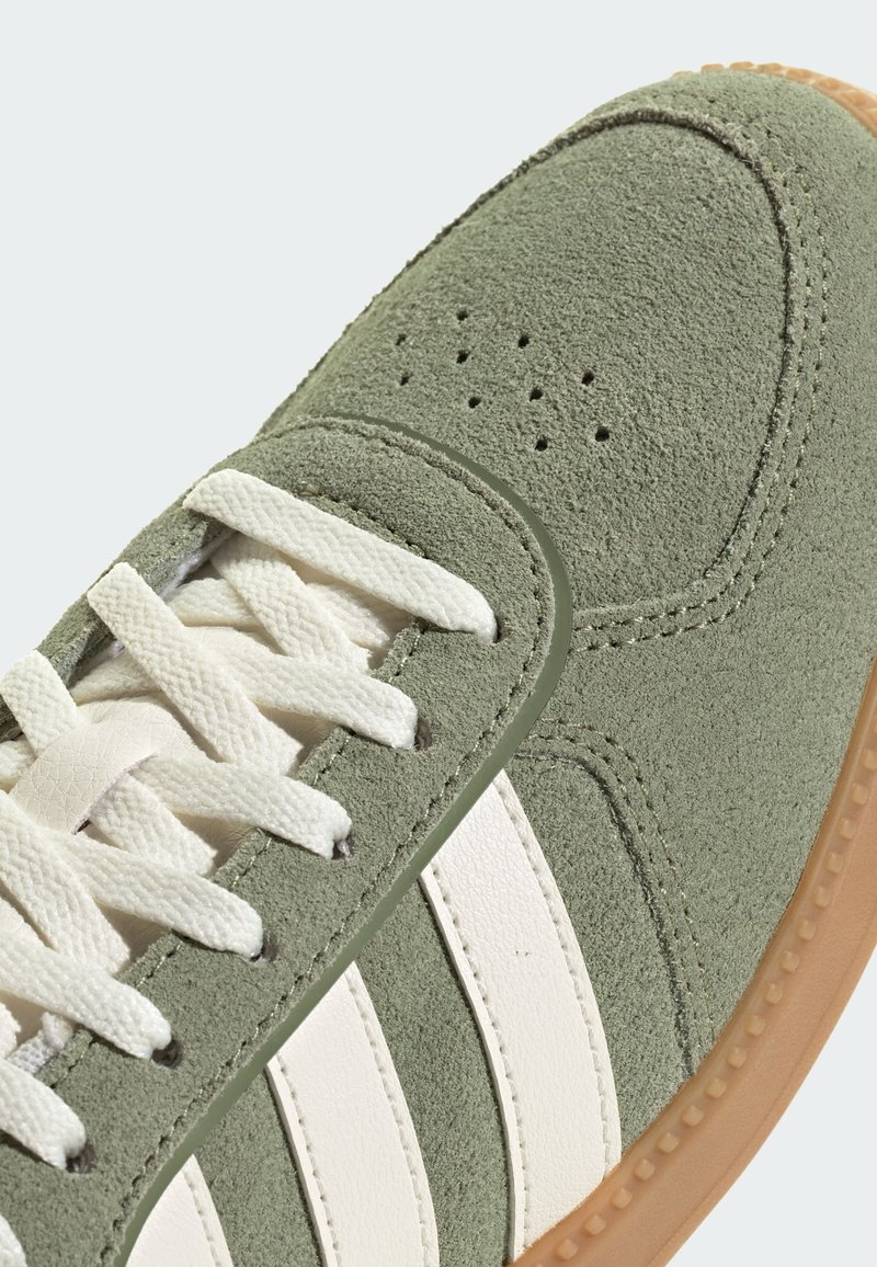 Grüner Wildledersneaker mit weißen Ledersstreifen, strukturiertem Obermaterial, rundem Zehenbereich und flachen weißen Schnürsenkeln. Gummisohle in Karamellfarbe.