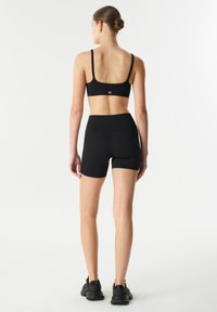 Zwarte sportbeha met dunne bandjes en logo, gecombineerd met zwarte hooggetailleerde shorts. Het materiaal lijkt glad en rekbaar.