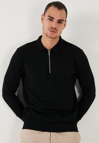 Sudadera negra con cierre de cremallera y cuello tipo polo, de mangas largas; presenta un tejido texturizado y un detalle de cremallera plateada visible.