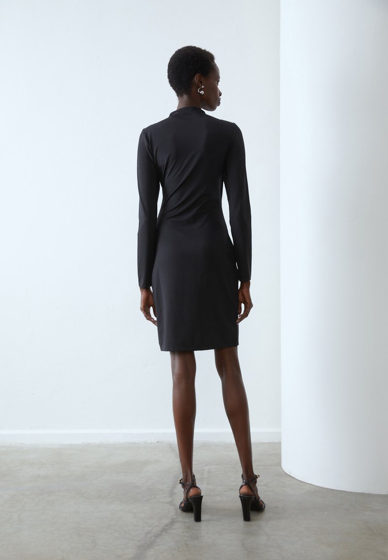 Abito lungo nero a maniche lunghe realizzato in tessuto liscio, silhouette aderente, lunghezza al ginocchio, con collo alto e design minimalista.