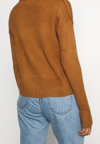 Pull marron tricoté avec un ourlet et des poignets côtelés, offrant une coupe ample. Associé à un jean taille haute bleu clair, vue de dos.