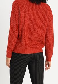 Pull rouge tricoté avec un motif texturé, coupe ample et ourlet côtelé. Associé à un pantalon noir à rayures fines.
