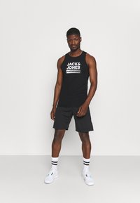 Débardeur noir sans manches avec le logo blanc "JACK & JONES PERFORMANCE", associé à un short noir et à des baskets blanches avec des accents bleus.