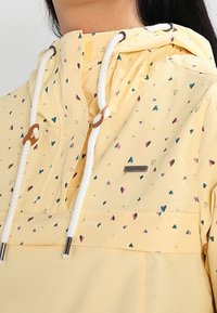 Gul huva-jacka med små flerfärgade hjärt-mönster, vita snörningar med bruna stoppare, bärs av en person som visas från hals till bröst.