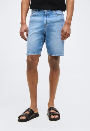 Mężczyzna w jasnoniebieskich szortach dżinsowych, czarnym bezrękawniku i czarnych sandałach wsuwanych stoi na białym tle.