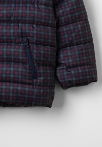 Giaccone imbottito con un motivo a scacchi blu navy e bordeaux. Presenta una tasca con zip e un tessuto testurizzato con un design trapuntato.