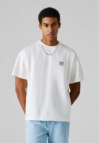 Λευκό βαμβακερό T-shirt με κοντά μανίκια, με τετράγωνη κοπή, που διαθέτει ραμμένη ετικέτα με την επιγραφή "Red Bull Racing" στο στήθος. Φορεμένο με ανοιχτόχρωμο μπλε τζιν παντελόνι.