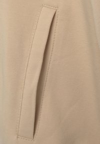 Beige fabric with a smooth texture features a side slit pocket, outlined with subtle stitching, emphasizing a simple and functional design.