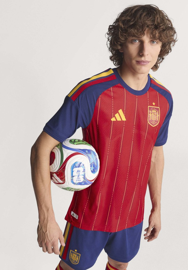 Maglietta da calcio rossa e blu con strisce verticali, dettagli gialli e logo; tiene in mano un pallone multicolore con un design che presenta stelle.