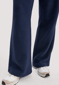 Benen gekleed in marineblauwe wijde fleecebroeken met witte en beige sportschoenen op een lichte achtergrond.