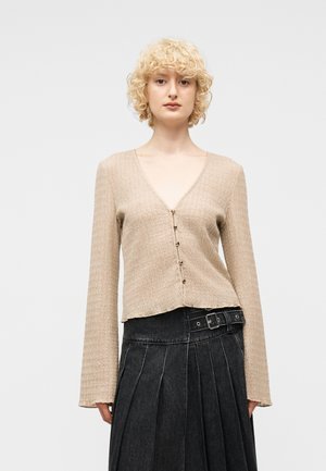 Blond osoba s kudrnatými vlasy nosící béžový texturovaný kardigan s výstřihem do V a knoflíky a černou vysokým pasem plisovanou sukni s páskem.