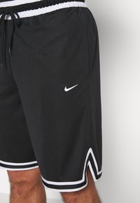 Shorts de sport noirs avec bordure blanche, logo Nike sur la cuisse gauche, taille élastique et cordon de serrage, portés sur la partie inférieure du corps d'une personne.