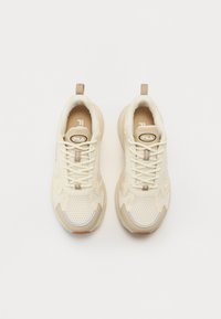 Zapatillas de running color crema con parte superior de malla, cuello acolchado y acentos beige. Presentan un diseño de cordones y un logotipo en la lengua. Suela de goma plana.