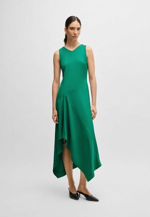 Green asymmetric dress in flowing fabric, featuring a sleeveless design, a V-neckline, and a draped hem. Worn with pointed black shoes.