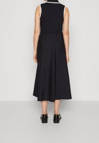 Kobieta stojąca, ubrana w czarną, bezrękawnikową sukienkę midi z dopasowanym pasem i czarnymi skórzanymi butami, odwrócona plecami na jednolitym tle.