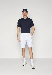 Μπλε σκούρο πόλο μπλουζάκι με κοντά μανίκια και γιακά, συνδυασμένο με λευκά σορτς. Ολοκληρώνεται με λευκά αθλητικά παπούτσια και μπλε σκούρο καπέλο.