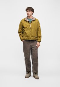 Νεαρός άνδρας που φοράει μουσταρδί ζακέτα με φερμουάρ, πράσινο πουκάμισο, γκρι παντελόνι και γκρι μπότες, στέκεται με τα χέρια στις τσέπες σε λευκό φόντο.