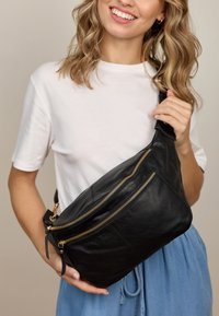Bolso cruzado de cuero negro con dos bolsillos con cremallera dorada, textura suave y forma triangular, llevado sobre una camiseta gris claro.