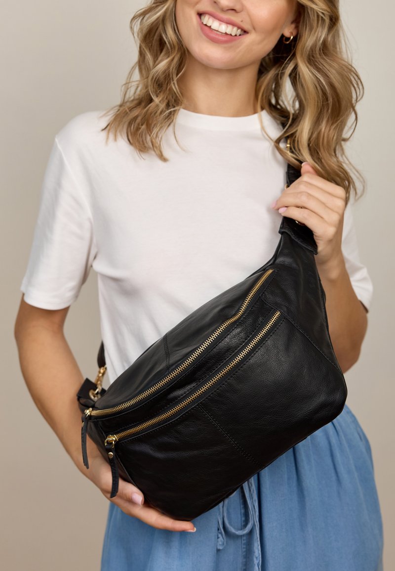 Bolso cruzado de cuero negro con dos bolsillos con cremallera dorada, textura suave y forma triangular, llevado sobre una camiseta gris claro.