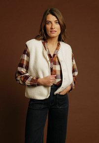 Gilet en polaire blanc avec des boutons-pression porté par-dessus une chemise à carreaux marron et crème. Le mannequin se tient devant un fond brun. Un jean noir est visible.