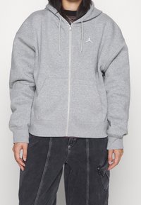 Sudadera gris con cremallera y bolsillo canguro, cordones, puños y cintura acanalados. Presenta un pequeño logo blanco en el lado izquierdo del pecho.