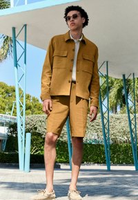 Chaqueta de lino mostaza con dos bolsillos en el pecho, combinada con pantalones cortos a juego. El modelo lleva zapatos de color claro y gafas de sol redondas.