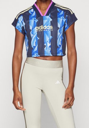 Personne portant un haut de sport court Adidas bleu et noir avec des leggings beige taille haute, debout devant un fond uni.