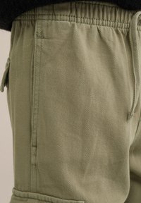 Gai zaļi kravas bikses ar teksturētu virsmu, kas aprīkotas ar elastīgu jostasvietu un šnori, sānu kabatām un smalkiem šuvju detalizējumiem.