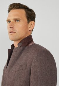 Giacca marrone a motivo Herringbone con colletto alto e patta con bottoni. Realizzata in tessuto testurizzato, presenta una tasca sul petto.
