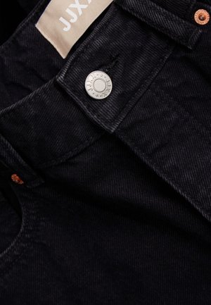 Schwarze Denim-Jeans mit glatter Oberfläche, ausgestattet mit einem silbernen Knopfverschluss, kupfernen Nieten und einem Stoffetikett mit "JJX"-Markenlogo.