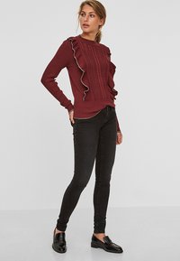 Suéter de punto burdeos con detalle de volantes a lo largo de un lado, patrón texturizado y diseño ajustado. Combinado con jeans ajustados negros y mocasines.