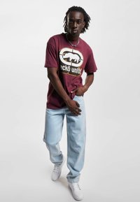 Mørkebrun bomulls t-skjorte med svart og hvit grafisk trykk, kombinert med lyseblå denimjeans og hvite sneakers, satt mot en hvit bakgrunn.