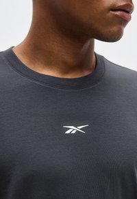 Κοντινό πλάνο ενός ατόμου που φοράει σκούρο γκρι μπλουζάκι με στρογγυλή λαιμόκοψη και μικρό λευκό λογότυπο Reebok στο κέντρο του στήθους.