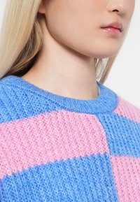 Posnetek bliže spodnjega dela obraza in vratu osebe, ki nosi modro in roza pleteno pulover s rebrastim vzorcem.