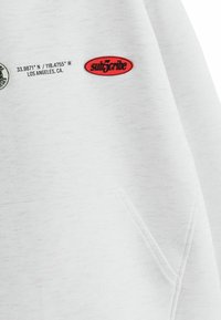 Sweat-shirt blanc avec une surface texturée. Elle présente un écusson ovale rouge avec "sub5cribe" et des coordonnées imprimées ainsi que des détails de localisation.