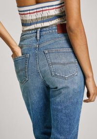 Jeans in denim blu a vita alta con gamba affusolata, tasche posteriori distintive e una toppa in pelle marrone del marchio sulla vita.