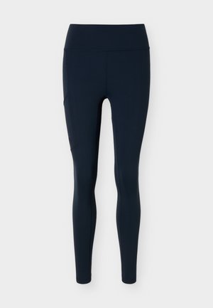 Tmavomodré dámske legíny s vysokým pásom, bočným vreckom, priliehavým strihom a dĺžkou po členky na svetlosivom pozadí.