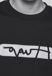 Sudadera negra con un acento rectangular blanco y letras negras bordadas. La tela parece suave con una textura lisa.