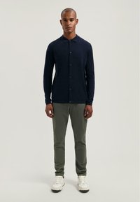 Námorně modrá polo košile s dlouhým rukávem, límcem a zapínáním na knoflíky, kombinovaná s olivově zelenými kalhotami a bílými teniskami.