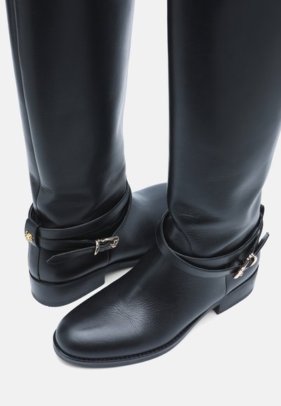 Paire de bottes noires en cuir montantes jusqu'aux genoux avec talons bas et sangles décoratives à la cheville avec boucles métalliques sur fond blanc.