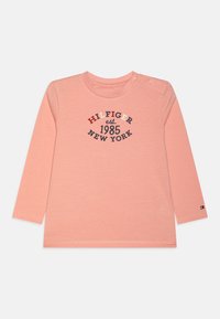 Långärmad, ljusrosa t-shirt med rund halsringning, pryds av marinblå och röda tryckta texter: "HILFIGER est. 1985 NEW YORK," och en liten logotyp på ärmen.